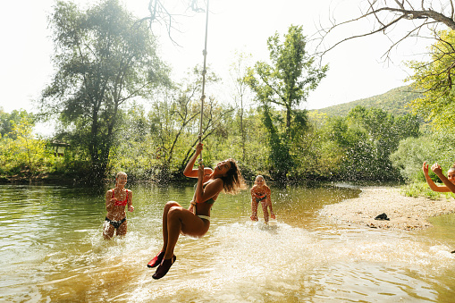 Photo of friends enjoying a scorching summer day by the river, joyfully swinging on a rope resembling liana.