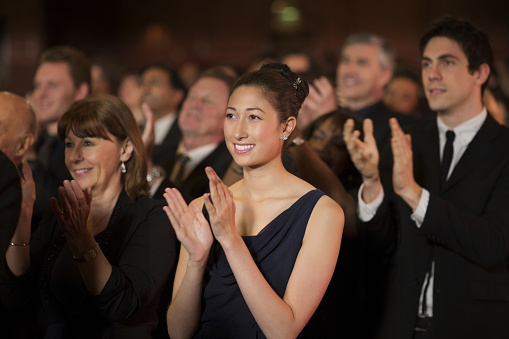 Side view of smiling spectators clapping hands in opera house. Men and women are watching theatrical performance. They are in elegant wear.