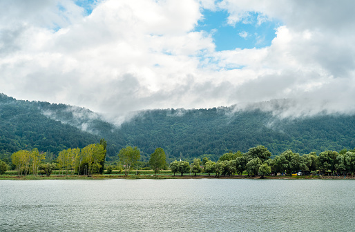 Mountain landscape with a lake, trees, surrounded by moving clouds.