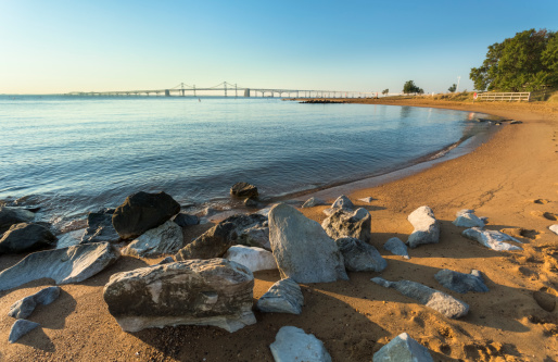 The early morning sun casts shadows of the large stones on the golden sands of Sandy Point State Park as the Chesapeake Bay Bridge stretches out along the horizon.