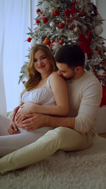 A man tenderly kisses his pregnant wife celebrating the New Year near the Christmas tree. The concept of New Year and Christmas holidays