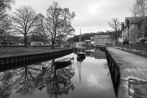 Black & white image of the River Dart, that runs through the town of Totnes, in South Devon. Historic Wharf buildings stand tall by the harbour, where boats are moored. The reflection of the nearby trees are defined in the water.