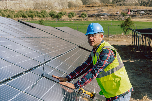 An electric engineering mature professional male with a blue helmet and worker vest is captured amid work at a photovoltaic farm, laptop in hand and drill. This image signifies the integration of technology and expertise in the maintenance, monitoring, or optimization of the solar panel energy production system. The engineer's use of a laptop suggests engagement in tasks related to data analysis, system monitoring, or perhaps planning for the efficient operation of the photovoltaic farm. This portrayal reflects the intersection of engineering and renewable energy technology in the contemporary pursuit of sustainable power solutions