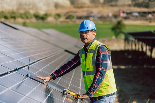 An electric engineering mature professional male with a blue helmet and worker west is captured amid work at a photovoltaic farm, laptop in hand and drill. This image signifies the integration of technology and expertise in the maintenance, monitoring, or optimization of the solar panel energy production system. The engineer's use of a laptop suggests engagement in tasks related to data analysis, system monitoring, or perhaps planning for the efficient operation of the photovoltaic farm. This portrayal reflects the intersection of engineering and renewable energy technology in the contemporary pursuit of sustainable power solutions