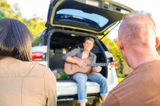Group of Man and woman friends playing guitar and singing together during camping on summer holiday vacation. Happy people relax and enjoy outdoor lifestyle with road trip travel at countryside.