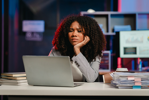 bored African American office worker with an afro, resting her cheek on her hand while looking at a laptop screen.