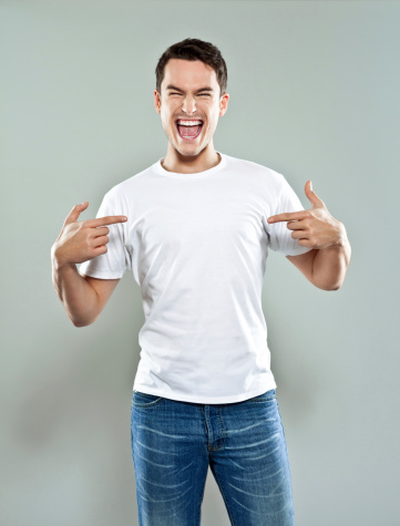 Portrait of happy young man wearing white t-shirt, pointing with fingers at his torso and laughing at camera. Studio shot, grey background.