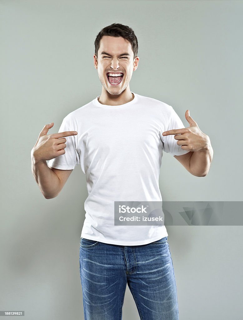 Excited young man Portrait of happy young man wearing white t-shirt, pointing with fingers at his torso and laughing at camera. Studio shot, grey background. Men Stock Photo