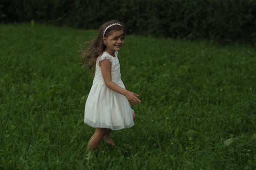A young girl runs joyously through a field, the young girl's white dress fluttering behind the young girl. image taps into the growing appreciation for simple, unplugged childhood moments in nature