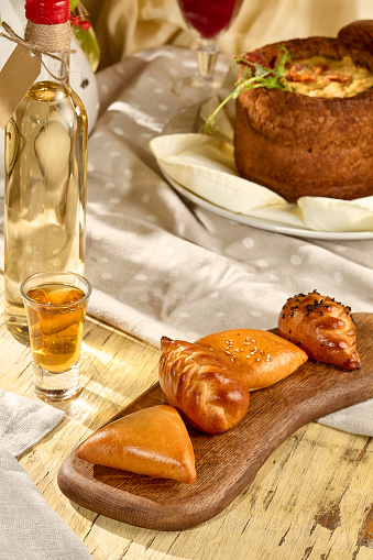 An assortment of pirozhki (pie) on a wooden board, with a shot of traditional drink.