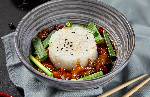 Aesthetic composition of Kung Pao chicken with rice in a grey bowl on a black textured table with chopsticks and textile. Ingredients surround the dish on a black background.