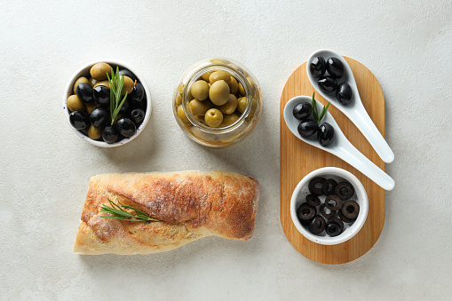 Olives in bowls and jar, baguette on white background, top view