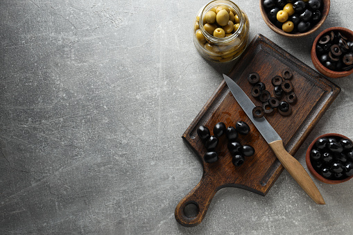 Olives in bowls and jar, kitchen board and knife on gray background, space for text