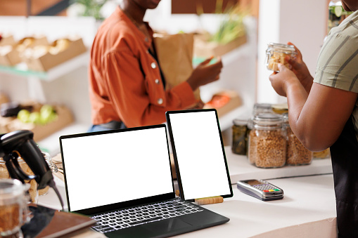 Digital screens on counter showing blank chromakey mockup template intended for eco friendly store advertisement. Tablet and laptop at cashier desk with isolated white screen.