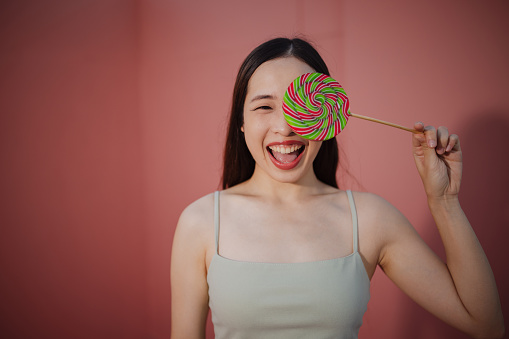 A joyful woman poses with a colorful spiral lollipop against a soft pink backdrop.