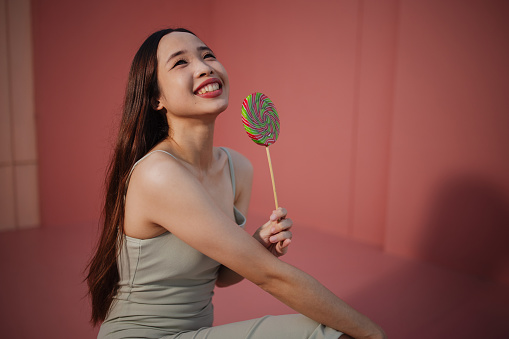 A joyful woman poses with a colorful spiral lollipop against a soft pink backdrop.