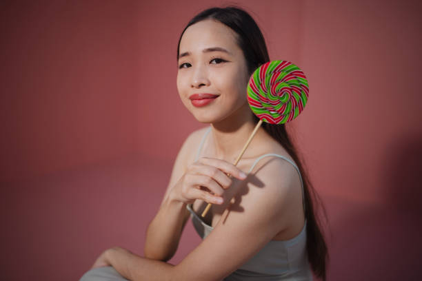 A joyful woman poses with a colorful spiral lollipop against a soft pink backdrop.