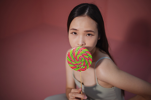 A woman poses with a colorful spiral lollipop against a soft pink backdrop.