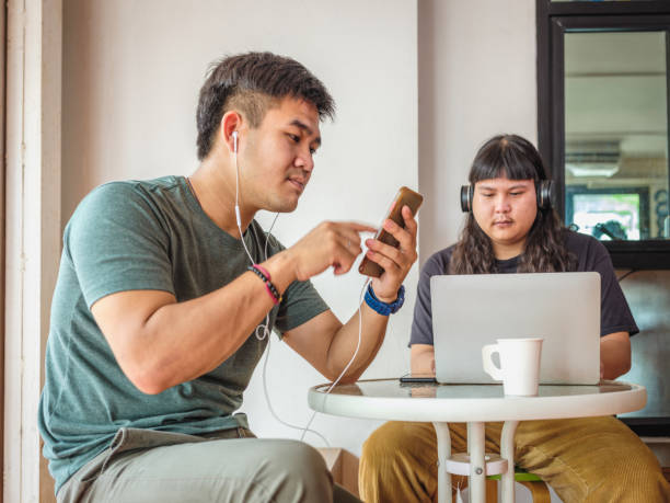 istock Two young Asian men using mobile phone and laptop computer while sitting at coffee shop cafe with skateboard 1881782277