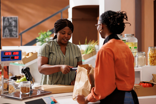 Female consumer paying with credit card at checkout counter, purchasing organic pantry staples at local supermarket. African american women at cashier desk using pos machine for seamless transactions.