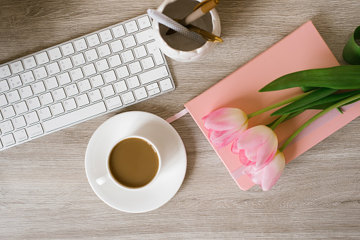Home office workplace: cup of coffee, notebook, keyboard, tulip flowers, brushes, on wooden background background