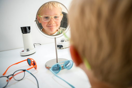 Enthusiastic young Caucasian boy testing frame fit, array of playful eyeglasses designs, cheerful optician assistance.