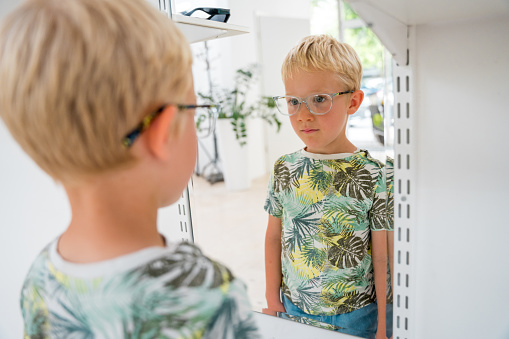 Caucasian young boy examines his reflection as he tries on a clear-framed pair of glasses, standing in a bright optometry store.