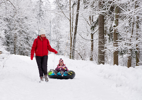 A young woman rides an 8-year-old Caucasian girl on an inflatable ring in the snow, on a walking path. The child is having fun.