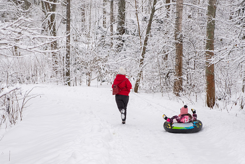A young woman rides an 8-year-old Caucasian girl on an inflatable ring in the snow, on a walking path.