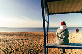 woman in an autumn jacket and hat is sitting alone on a bench on a deserted autumn beach on a sunny day