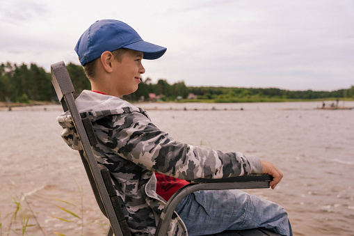 A school-age boy in solitude sits on a chair at the calm lake shore, reflecting on the serene beauty of nature, surrounded by peaceful tranquility and scenic landscapes.