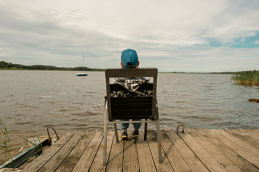 An unrecognizable school-age boy sits alone on a chair on the calm shore of the lake with his back to the camera, thinking and dreaming