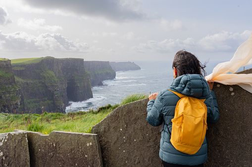 Unknown woman on her back with yellow backpack enjoying a splendid view from the Moher´s Cliff in Ireland with scarf fluttering in the wing