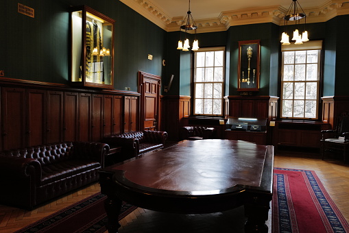 This image offers a view into the boardroom of Belfast City Hall, where the air of decision-making lingers in the dignified silence. The room is adorned with polished wooden panels and furnished with stately leather chairs, ready for the next meeting of minds. The light from the elegant pendant lamps casts a soft glow on the gleaming tabletop and the richly colored runner on the floor. Portraits and ceremonial maces displayed in glass cases add a touch of historical significance, encapsulating the weight of civic duty and the heritage that has shaped the city.