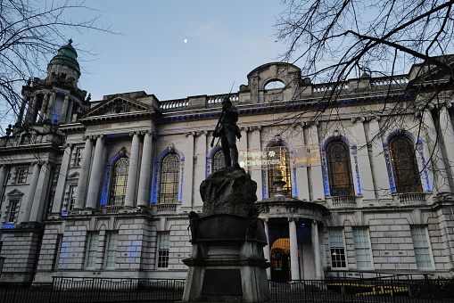 As dusk falls, Belfast City Hall is enlivened with the festive glow of holiday lights. This photograph captures the historic building adorned with the message 'Happy Christmas Belfast,' bringing seasonal cheer to all who pass by. A silhouette of a statue stands in the foreground, bearing witness to the city's seasonal transformation. The soft light of the moon above complements the blue-hued illuminations, creating a picture-perfect scene that celebrates the joy and community spirit of the holiday season.