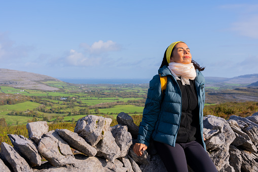 Latin woman enjoys an Irish landscape while resting leaning on a stone wall and taking a deep breath with the sea in the background