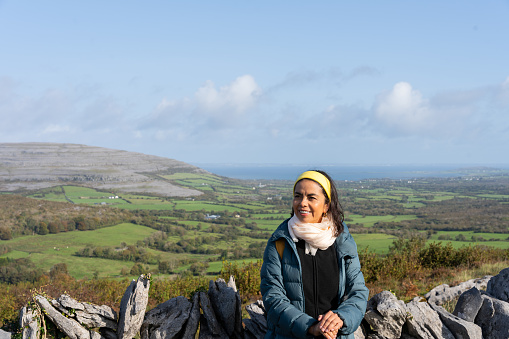 Latina female hiker with scarf enjoys beautiful scenery while resting against a stone wall in Ireland