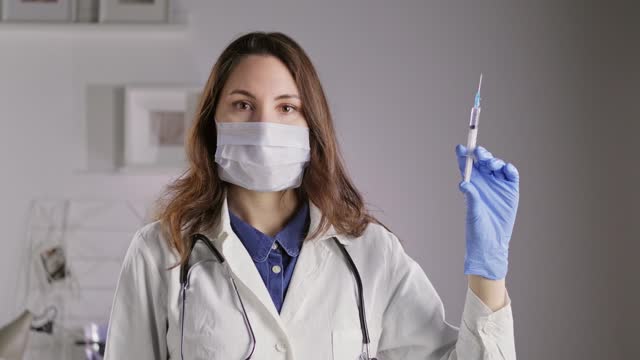 A female doctor in a mask and gloves looks into the camera, holds a syringe and sprays liquid out of it. Medicine and Pharmaceuticals