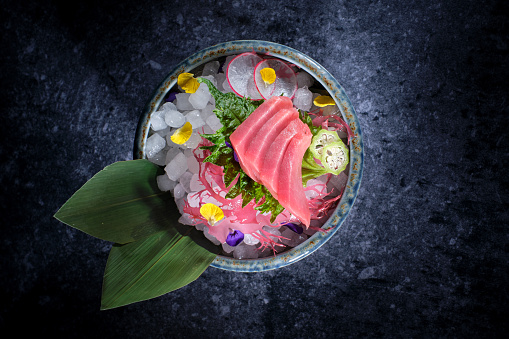 Top view of fresh tuna sashimi served in bowl with ice cubes and green banana leaves placed on gray background