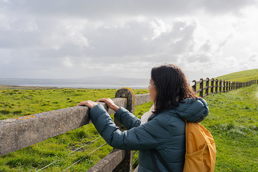 Latin woman with backpack leaning on a fence while enjoying a moment of rest from trekking in an Irish landscape