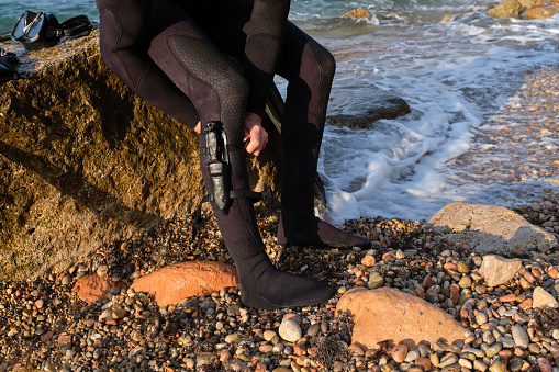 Crop anonymous male diver in black wetsuit sitting on boulder on shore of sea and tying knife to leg in daylight
