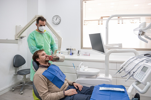A dentist taking an X-ray of a patient's mouth sitting in a dentist's chair with the instruments around him.
