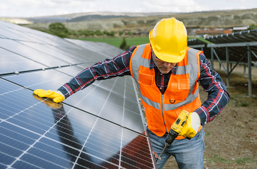 The synergy of technology and sunlight in this impactful, showcasing a technician engaged in the solar panel installation process. Safely harnessing solar power with a drill on the roof structure, the scene unfolds on a sunny day at a solar energy power station farm. The image captures the dedication and precision required for effective solar energy deployment. Ideal for conveying progress in renewable energy, this photo is perfect for energy-related promotions, environmental publications, and social media content championing a brighter and sustainable future. Solar panel installation, Technician, Harnessing, Solar power, Drill, Roof structure, Sunny day, Solar energy power station farm, Dedication, Precision, Progress, Energy-related promotions, Environmental publications, Social media, Sustainable future