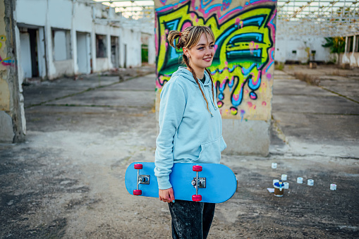 Smiling young woman with her skateboard standing in front of a wall with colorful graffiti