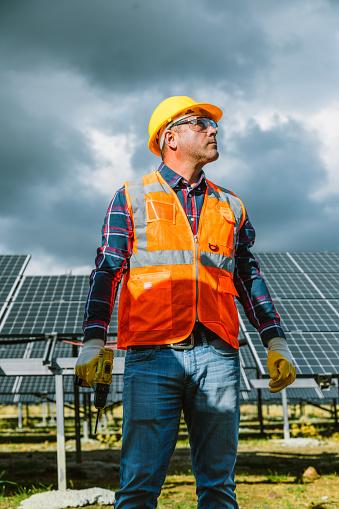 Portrait of a serious mid-adult worker wearing a yellow work helmet and reflective vest. The worker is holding an electrical screwdriver or drill, preparing to install solar panels in a natural sun energy photovoltaic farm power station. The image reflects the commitment and proficiency required for effective solar energy deployment. Ideal for conveying professionalism and dedication to clean energy, this photo is perfect for energy-related promotions, engineering publications, and social media content celebrating advancements in sustainable power. Mid-adult worker, Serious look, Yellow work helmet, Reflective vest, Electrical screwdriver, Drill, Solar panels, Photovoltaic farm, Power station, Expertise, Determination, Clean energy, Energy-related promotions, Engineering publications, Social media, Sustainable power