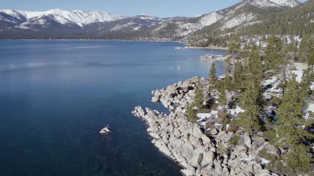 A low-flying, 4K drone shot over Lake Tahoe, California, during the winter season. The camera slowly moves forward, and rotates right around a small peninsula, along the lake's snow-covered coastline.