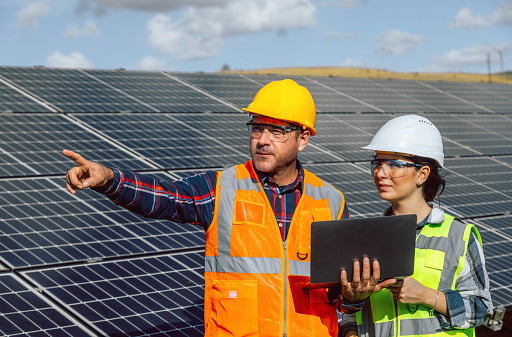 Young electrical engineer woman and business man standing in front of wind turbines checking and working about technical problems and writes the results of measurements with laptop pc in sun power plant electric energy station. xxxl