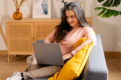 Mature woman working on laptop at home, sitting on sofa.