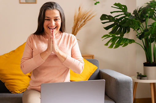 Cheerful mature woman sitting on sofa and using laptop at home