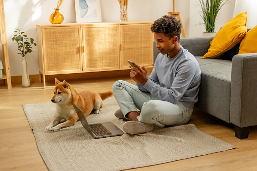 african american man using smartphone while sitting with shiba inu dog on carpet at home